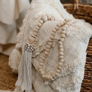 Può includere: Una coperta color crema e strutturata con una ghirlanda di perline di legno e nappa. La ghirlanda ha grandi perline di legno rotonde e un elemento decorativo all'estremità. La nappa è realizzata in filato morbido e chiaro.