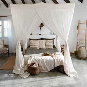 Puede incluir: Un dormitorio con una cama con dosel cubierta con tela blanca. La cama está hecha con ropa de cama gris y almohadas decorativas. Una cesta de mimbre y una bandeja de madera con fruta y decoración están al pie de la cama. Una escalera de madera y una linterna tejida también están en la habitación.