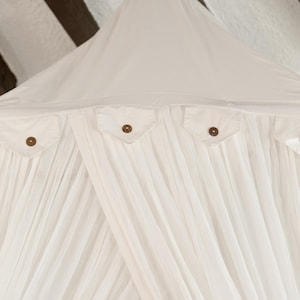 May include: A white canopy bed with a draped design. The canopy has four small pockets with wooden buttons. The fabric is gathered and flowing, creating a soft, airy aesthetic. The canopy is suspended from a white ceiling with dark brown wooden beams.