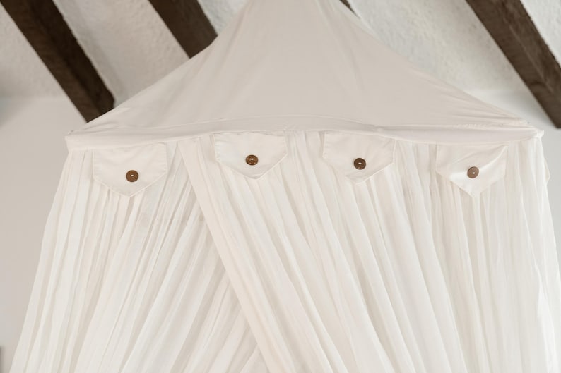 May include: A white canopy bed with a draped design. The canopy has four small pockets with wooden buttons. The fabric is gathered and flowing, creating a soft, airy aesthetic. The canopy is suspended from a white ceiling with dark brown wooden beams.