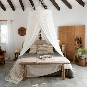 Op de afbeelding: Een slaapkamer met een hemelbed, met een witte luifel en een houten bank aan het voeteneinde van het bed. De kamer heeft een neutraal kleurenpalet met een vlechtwerk scherm en decoratieve kussens.