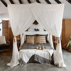 Puede incluir: Una cama con dosel con cortinas blancas transparentes sobre una colcha gris oscura. Cojines decorativos en tonos neutros y una manta con flecos añaden textura. Una bandeja de madera contiene objetos decorativos.