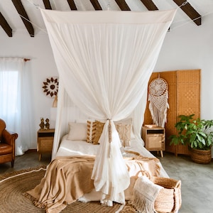 May include: A bedroom with a bohemian theme, featuring a bed draped with a sheer white canopy. The bed is adorned with decorative pillows and a tan throw blanket. A woven rug, a wicker screen, and a dreamcatcher add to the room's decor.