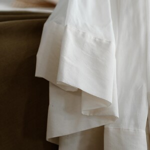 May include: Close-up of a white garment with wide, layered sleeves. The fabric appears to be a lightweight material, possibly linen or cotton, with a subtle texture. The sleeves have a contrasting, thicker hem.