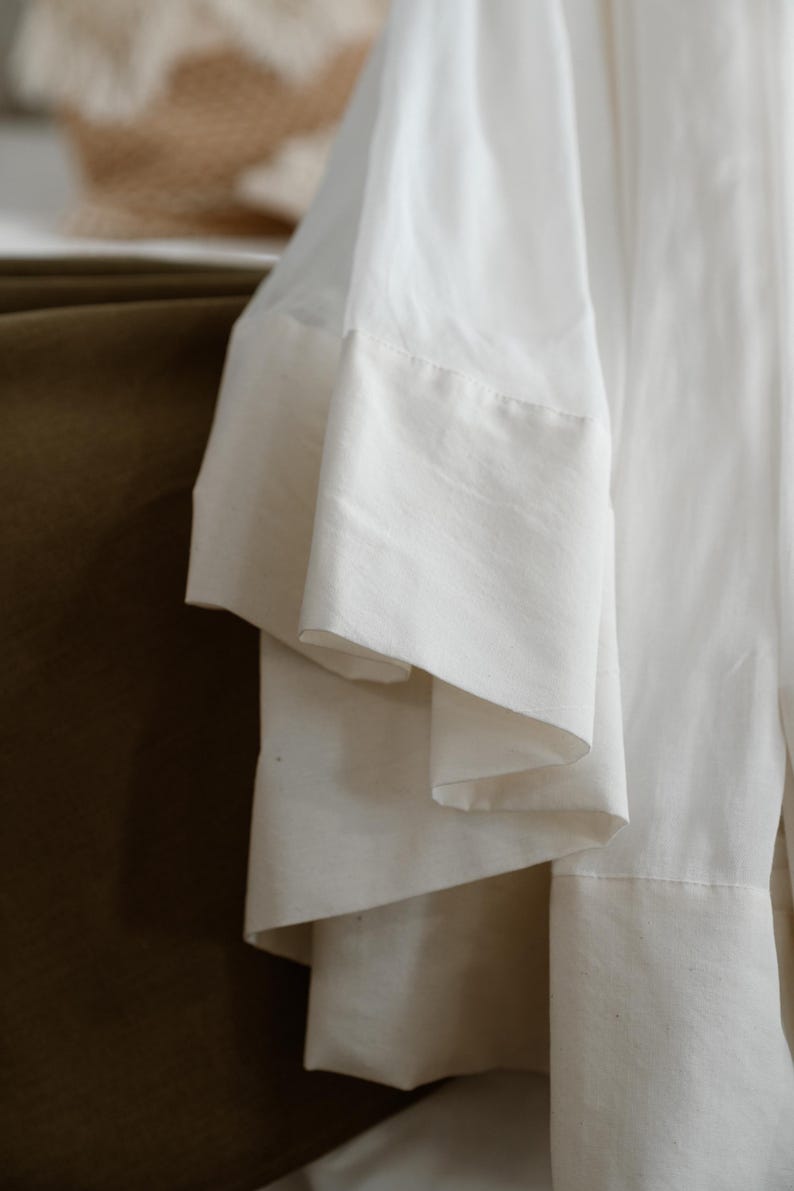 May include: Close-up of a white garment with wide, layered sleeves. The fabric appears to be a lightweight material, possibly linen or cotton, with a subtle texture. The sleeves have a contrasting, thicker hem.