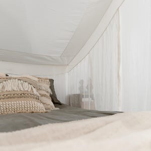 May include: A bed with a white canopy and a gray bedspread. Decorative pillows with textured, neutral-toned covers are arranged on the bed. A cream-colored blanket is draped over the foot of the bed.