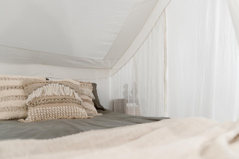 May include: A bed with a white canopy and a gray bedspread. Decorative pillows with textured, neutral-toned covers are arranged on the bed. A cream-colored blanket is draped over the foot of the bed.