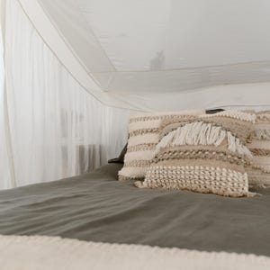 May include: A bed with a canopy and decorative pillows. The bed has a dark green sheet and a cream-colored blanket. The pillows are cream and beige with textured patterns and fringe.