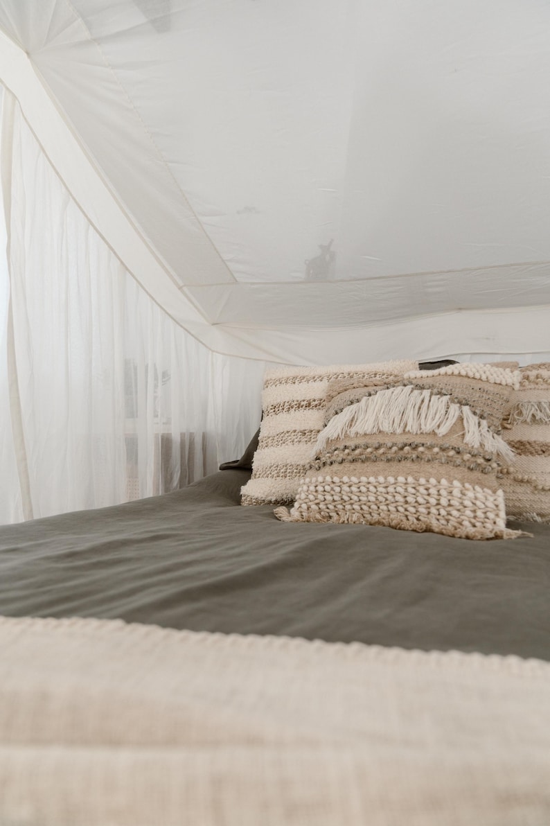 May include: A bed with a canopy and decorative pillows. The bed has a dark green sheet and a cream-colored blanket. The pillows are cream and beige with textured patterns and fringe.