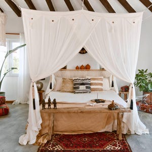 Puede incluir: Dormitorio con cama con dosel, con cortinas blancas transparentes y cabecero de madera. Cojines decorativos y un banco de madera con decoración se encuentran al pie de la cama. Una alfombra estampada y plantas completan el estilo bohemio.