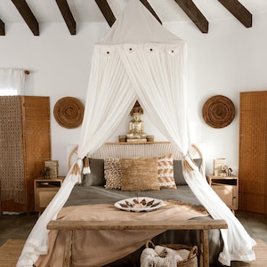 May include: A bedroom with a canopy bed, featuring a white draped canopy and a wooden bench at the foot of the bed. The room has a neutral color palette with woven accents, including a rug, pillows, and decorative wall hangings.