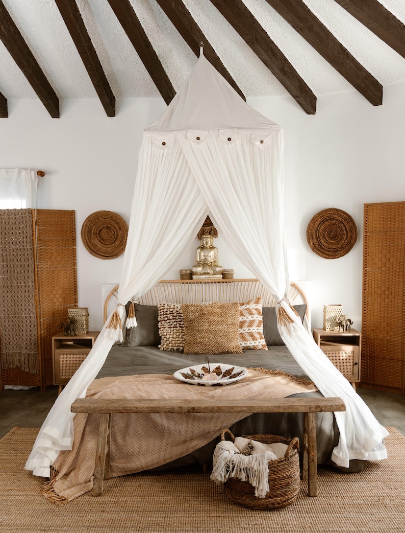 May include: A bedroom with a canopy bed, featuring a white draped canopy and a wooden bench at the foot of the bed. The room has a neutral color palette with woven accents, including a rug, pillows, and decorative wall hangings.