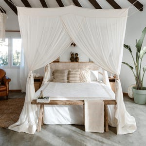 Puede incluir: Una cama con dosel con ropa de cama y almohadas blancas. La cama tiene un banco de madera a los pies y está cubierta con tela blanca transparente. Cojines decorativos y un cabecero tejido añaden textura. Una planta en maceta y mesitas de noche completan el ambiente.