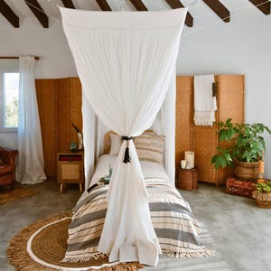 May include: A bedroom scene featuring a bed with a white canopy and a striped blanket. A round, fringed rug sits on the floor. Woven screens and a potted plant add to the bohemian aesthetic. The room has a neutral color palette with natural textures.