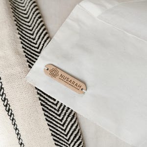 May include: Close-up of a textile with a black and white chevron pattern and a cream-colored fabric with black vertical stripes. A white fabric is in the background with a wooden tag that reads "NUSARAH".