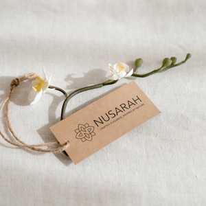 May include: A brown rectangular tag with the word "NUSARAH" and a floral design, attached to a natural twine string. The tag is accompanied by a sprig of white orchids with yellow centers, set against a white textured background.