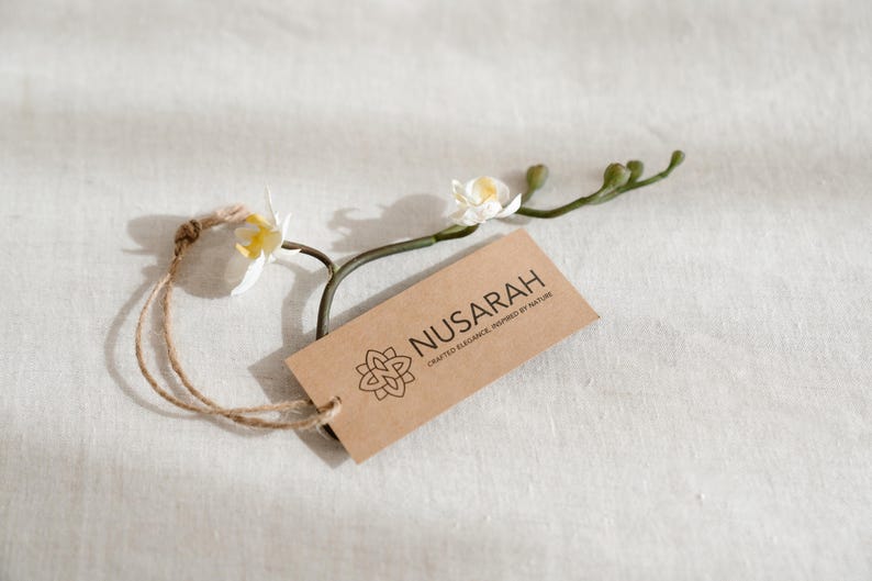 May include: A brown rectangular tag with the word "NUSARAH" and a floral design, attached to a natural twine string. The tag is accompanied by a sprig of white orchids with yellow centers, set against a white textured background.