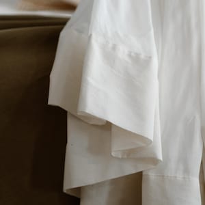 May include: Close-up of a white garment with wide, layered sleeves. The fabric appears to be a lightweight material, possibly linen or cotton, with a subtle texture. The sleeves have a contrasting, thicker hem.