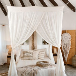 Puede incluir: Una cama con dosel con cortinas blancas y vaporosas. La cama está vestida con almohadas de tonos neutros y una cesta tejida. Una manta estampada cubre la cama. Un atrapasueños cuelga de una pantalla tejida.