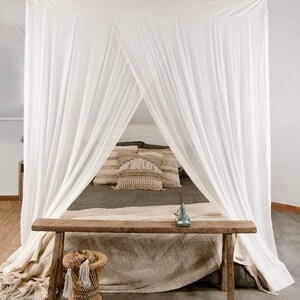 May include: A bedroom scene featuring a canopy bed with white drapes. A wooden bench sits at the foot of the bed, and a small woven stool is in the foreground. Decorative pillows and a blanket add texture to the bed.