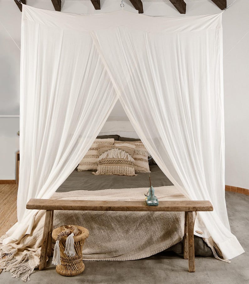 May include: A bedroom scene featuring a canopy bed with white drapes. A wooden bench sits at the foot of the bed, and a small woven stool is in the foreground. Decorative pillows and a blanket add texture to the bed.