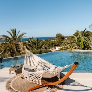 May include: A relaxing outdoor scene featuring a white hammock with decorative pillows, set near a pool. The hammock is suspended on a wooden frame, resting on a round jute rug. Palm trees and a clear blue sky complete the tropical vibe.