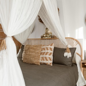 May include: A bedroom scene featuring a bed with a woven headboard and a draped canopy. The bed is made with a dark gray duvet and decorative pillows in natural tones. A gold Buddha statue sits on the headboard.