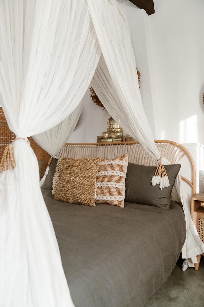 May include: A bedroom scene featuring a bed with a woven headboard and a draped canopy. The bed is made with a dark gray duvet and decorative pillows in natural tones. A gold Buddha statue sits on the headboard.