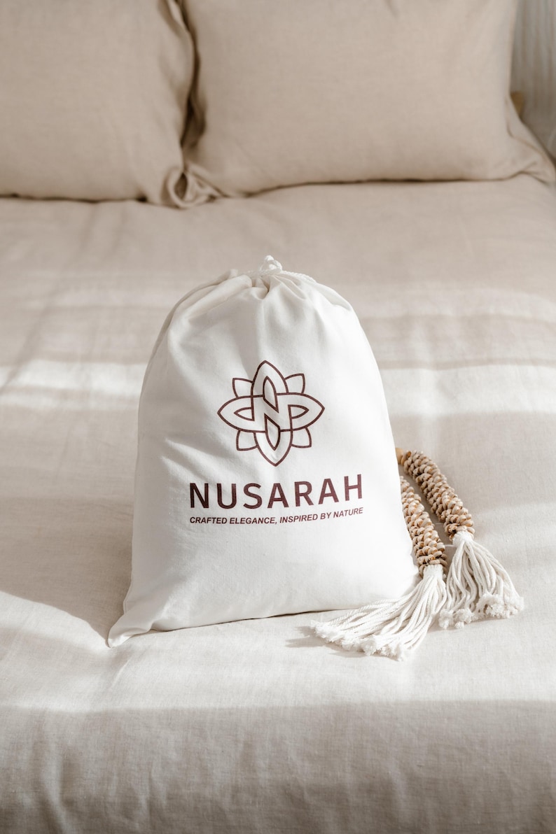 May include: A white drawstring bag with the brand name "NUSARAH" and a floral logo in brown. The bag is on a bed with beige linens, and a decorative tassel is next to it.