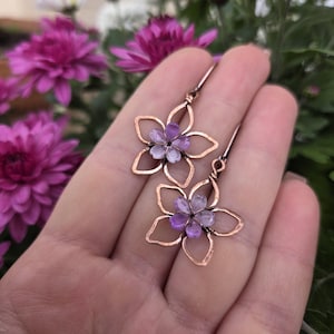 May include: Copper-colored flower earrings with purple and white gemstone centers. The earrings are held in a hand, with a background of pink flowers. The earrings have a delicate, openwork design.