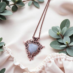 Può includere: Una collana con pendente avvolto in filo di rame con una pietra centrale azzurra. Il pendente ha una forma triangolare con intricati dettagli in filo. La catena della collana è anch'essa color rame ed è esposta su un tessuto color crema con fogliame verde.