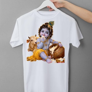 May include: A white t-shirt featuring a graphic print of a blue-skinned child with curly dark hair, wearing gold jewelry and a yellow garment. The child is sitting next to a small cow and a brown pot filled with white food, holding a piece of food.