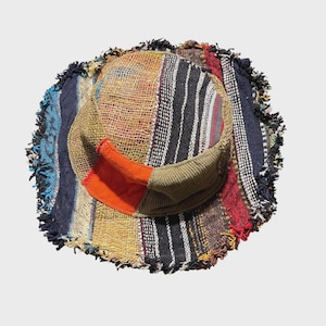 May include: A bucket hat constructed from various patchwork fabric strips in shades of blue, red, black, and tan. The hat features a frayed, uneven brim and a textured, woven appearance with a prominent bright orange rectangular fabric patch.