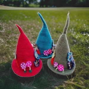 May include: Three pointed felt hats in red, blue, and olive green. Each hat features decorative felt appliques including small pink and purple mushrooms with white spots, and various leaf-shaped accents attached to the base of the cone.