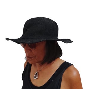 May include: A black textured hat with a wide, slightly wavy brim and a rounded crown. The hat is constructed from a woven material with a visible, coarse grain. The brim extends outwards from the base of the crown, providing a structured silhouette.
