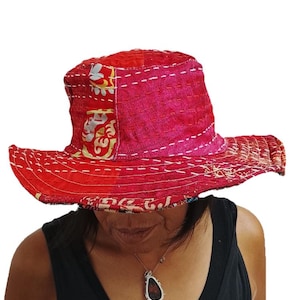 May include: A bucket hat crafted from patchwork fabric in shades of red and pink, featuring visible white running stitch embroidery throughout the crown and wide brim. The textile displays a mix of solid colours and subtle floral patterns.