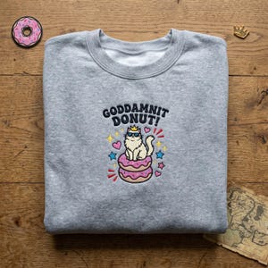 May include: A heather grey crewneck sweatshirt featuring an embroidered graphic of a white cat wearing a gold crown and blue sunglasses, sitting on two pink frosted doughnuts with sprinkles. The text reads GODDAMNIT DONUT! with colourful stars and hearts.