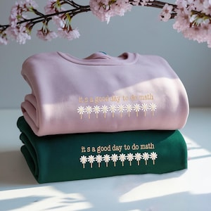 May include: Two sweatshirts, one light pink and one dark green, stacked on top of each other. The text "it's a good day to do math" is embroidered on each, along with a row of daisy flowers. The sweatshirts are folded neatly.