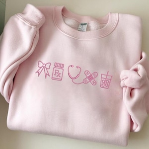 Puede incluir: Sudadera rosa claro con iconos médicos rosas bordados, incluyendo un lazo, un frasco de medicamentos, un estetoscopio, una venda y una bebida. La sudadera tiene cuello redondo y puños acanalados.