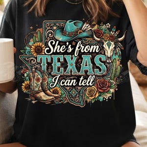 Pode incluir: Uma t-shirt preta com uma estampa gráfica com o texto "She's from TEXAS I can tell". O design inclui um chapéu de cowboy turquesa, um par de botas de cowboy florais, um crânio de boi, girassóis, rosas e cactos em formato de estado.
