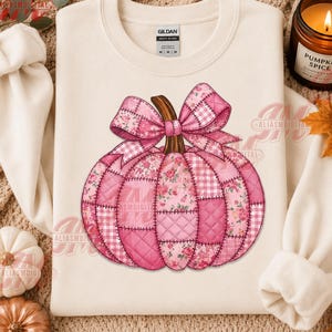 May include: A cream-colored crewneck sweatshirt featuring a large graphic of a pumpkin made from various pink quilted fabric patterns, including floral and gingham prints, topped with a large pink bow and a brown stem, with a Gildan brand label.