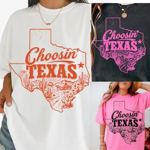 May include: Three t-shirts with a Texas theme. One white t-shirt with an orange graphic of the state of Texas, the words "Choosin' TEXAS", and a desert scene. A black t-shirt with a pink graphic and a pink t-shirt with a black graphic.