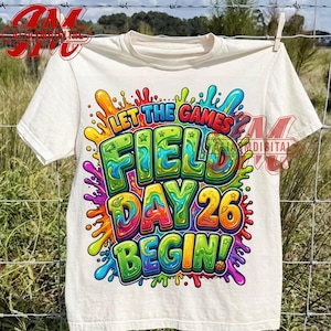 May include: A white t-shirt with a colorful graphic design. The design features the text "LET THE GAMES FIELD DAY 26 BEGIN!" in a vibrant, multi-colored, cartoon-style font with splashes of paint.