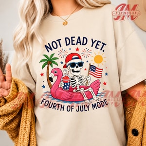 Peut inclure: Un t-shirt beige avec un motif de squelette portant des lunettes de soleil et une casquette rouge, assis dans une bouée flamant rose. Le squelette tient une boisson et un drapeau américain. Texte : NOT DEAD YET, FOURTH OF JULY MODE.