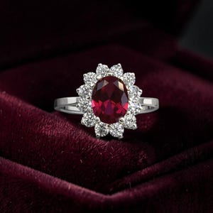 Puede incluir: Un anillo plateado con una gema roja oscura de corte ovalado, rodeada por un halo de piedras claras más pequeñas. El anillo se exhibe sobre un fondo de terciopelo burdeos oscuro, resaltando el brillo de las piedras.