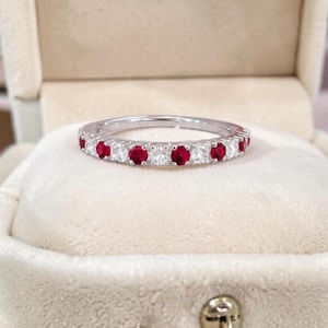 Könnte beinhalten: Ein silberner Ring mit abwechselnd roten und klaren Edelsteinen. Der Ring wird auf einer weißen, samtigen Oberfläche präsentiert. Die Edelsteine sind rund und leuchtend. Ein elegantes Schmuckstück.