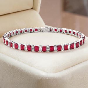 Pode incluir: Uma pulseira prateada com pedras preciosas vermelhas retangulares e pequenas pedras transparentes. A pulseira é exibida em uma caixa de joias creme. As pedras preciosas são espaçadas uniformemente.