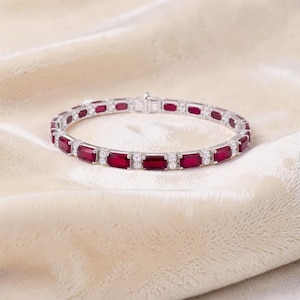 Puede incluir: Una pulsera plateada con piedras preciosas rectangulares de color rojo intenso y pequeñas piedras claras. La pulsera está colocada sobre una superficie texturizada de color crema. El cierre es visible y la pulsera es circular.