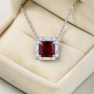 May include: A silver necklace with a square, deep red gemstone pendant. The pendant is surrounded by a halo of smaller, clear gemstones. The necklace is displayed in a beige jewelry box.