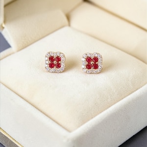 May include: A pair of gold-toned stud earrings, each featuring a square design with a cluster of red gemstones at the center, surrounded by a halo of smaller, clear stones. The earrings are presented in a white jewelry box.